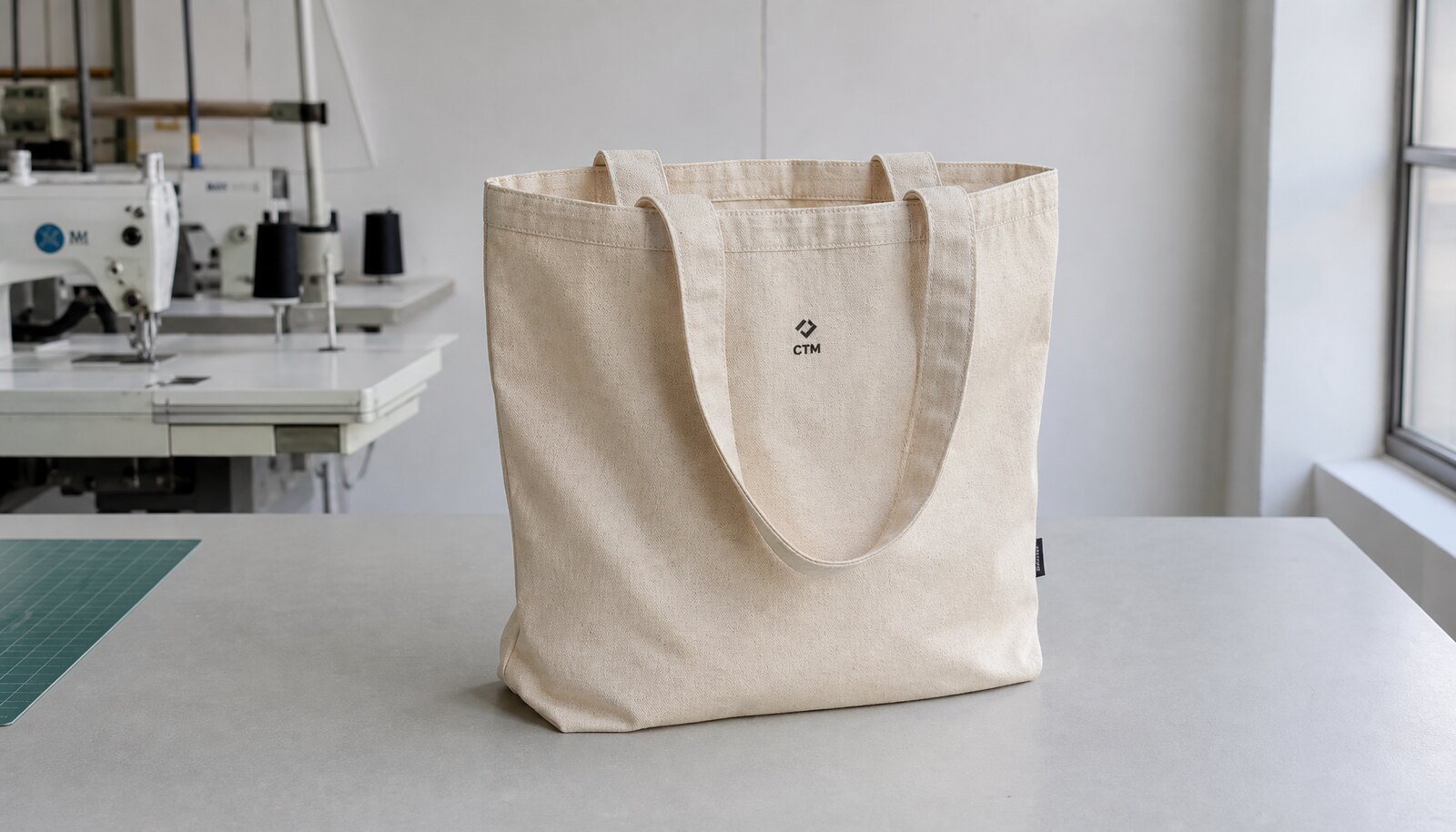 Natural canvas tote bag with a small CTM logo print and sewn side label displayed in a clean factory sample room