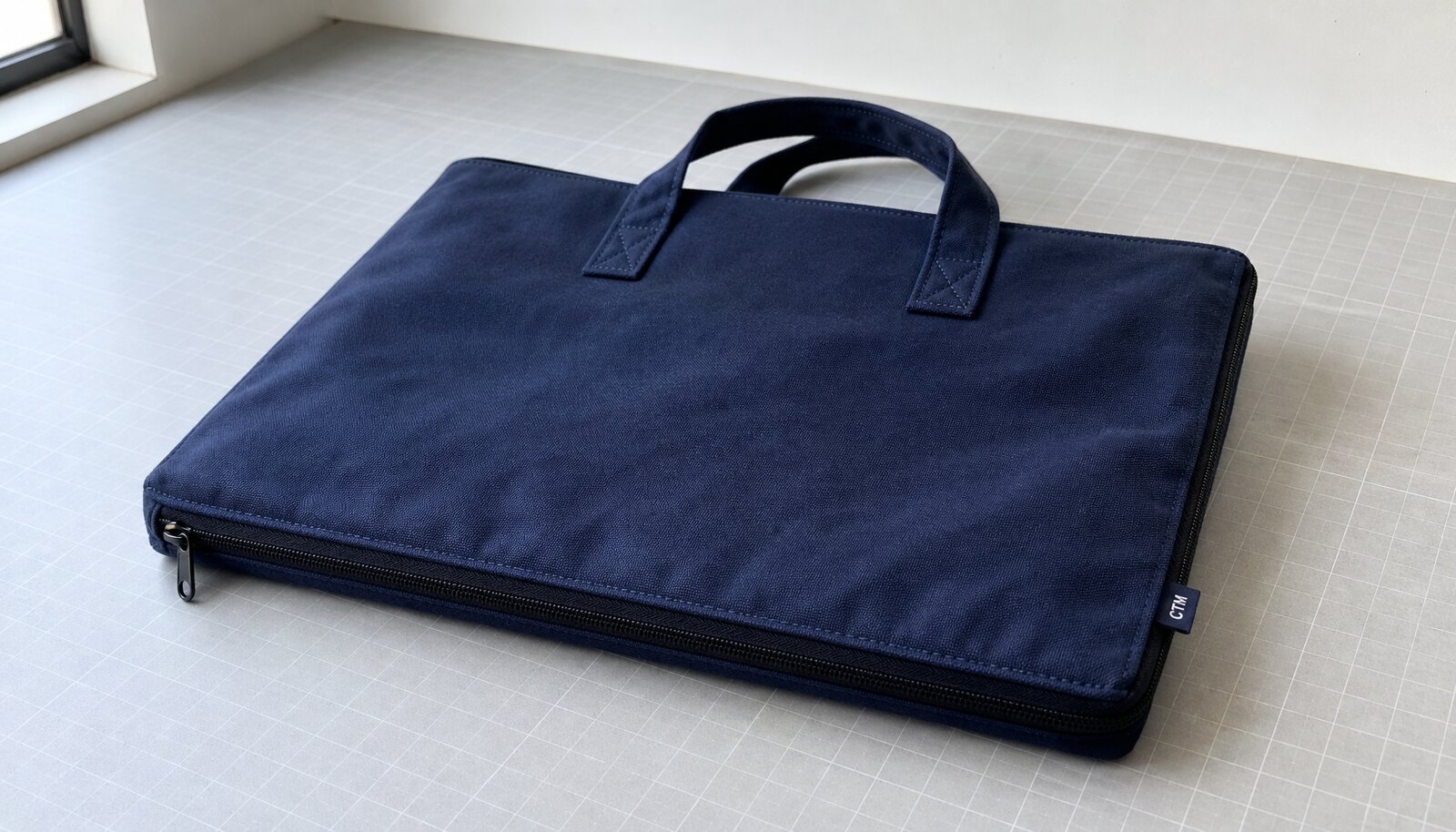 Navy canvas zipper portfolio with a woven CTM side label in a factory sample room.