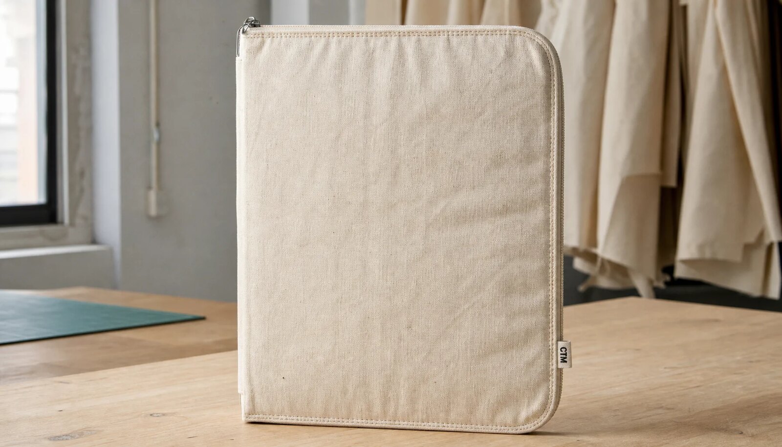 Natural cotton canvas zipper portfolio with visible stitching, matte zipper puller, and sewn CTM side label in a factory sample room.