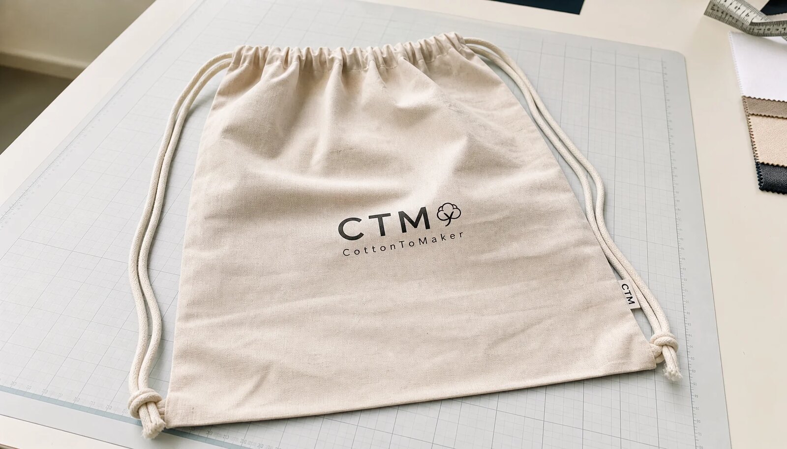 Natural cotton drawstring backpack with a simple CTM logo printed on the front and a small woven side label in a factory sample room.