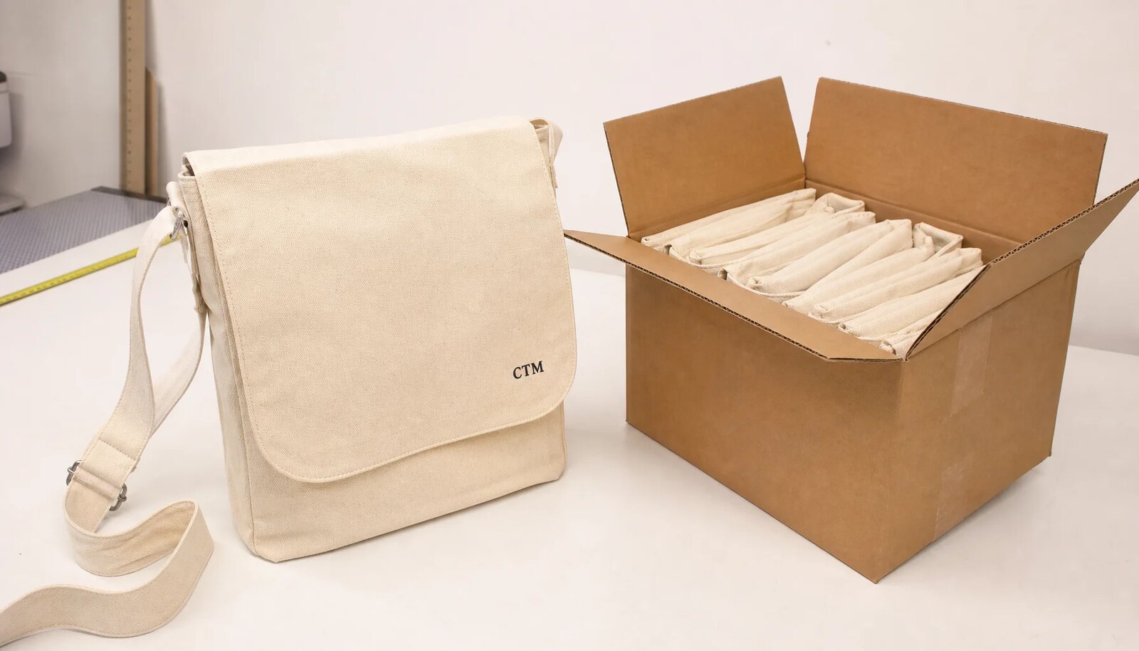 Custom canvas messenger bag with CTM logo beside an export carton for craft fair packing review