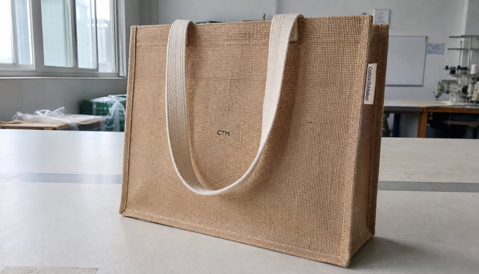 Natural jute tote bag with a small CTM logo and woven CottonToMaker side label in a sample room