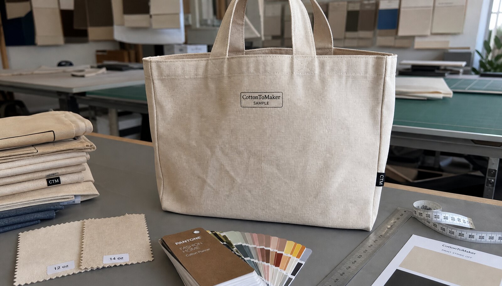 Natural heavy canvas tote bag with sample logo print, fabric swatches, ruler, and Pantone book on a factory sample table