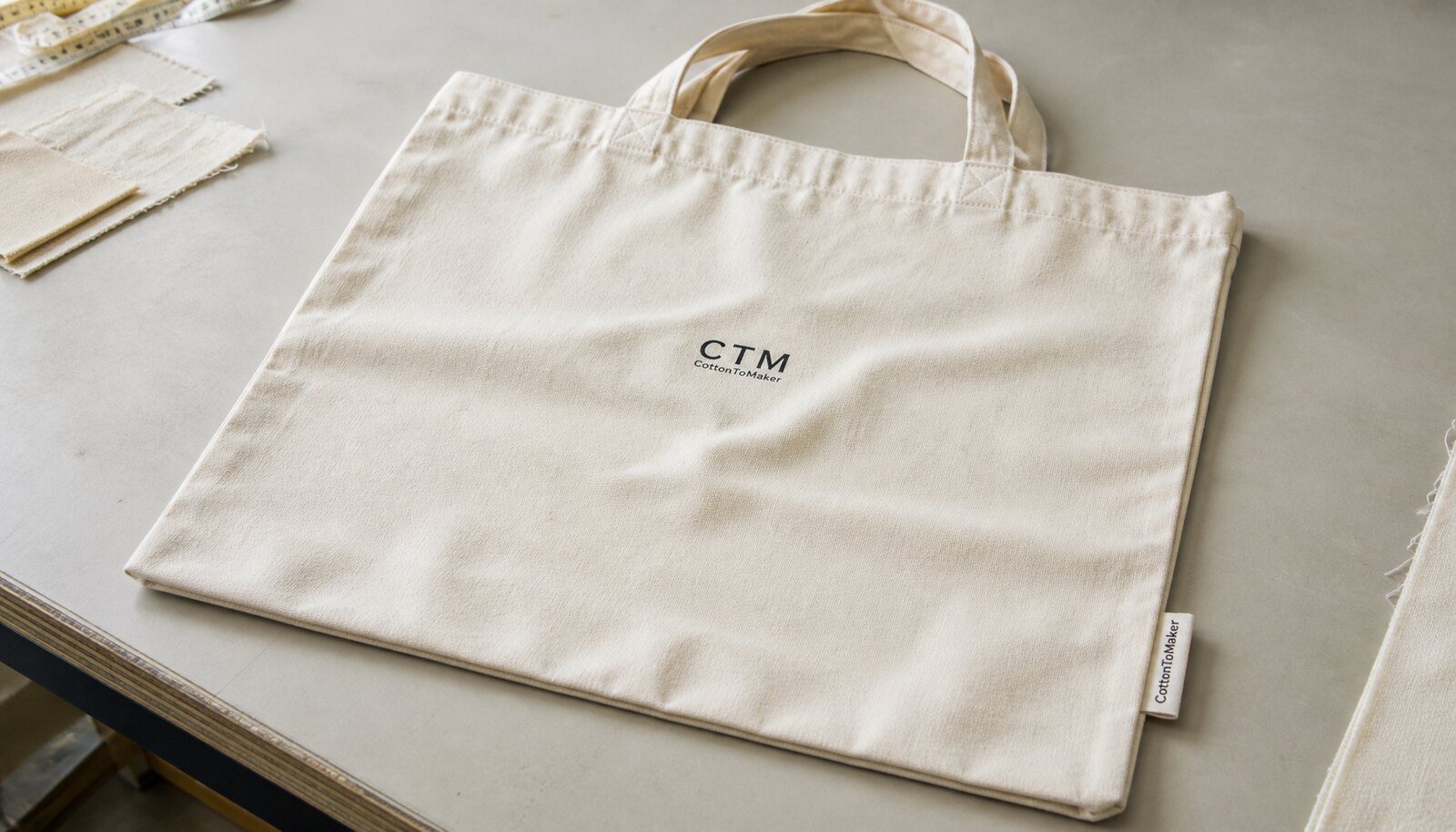 Natural canvas shopping bag with a simple CTM logo printed on the front and a CottonToMaker side label in a factory sample room