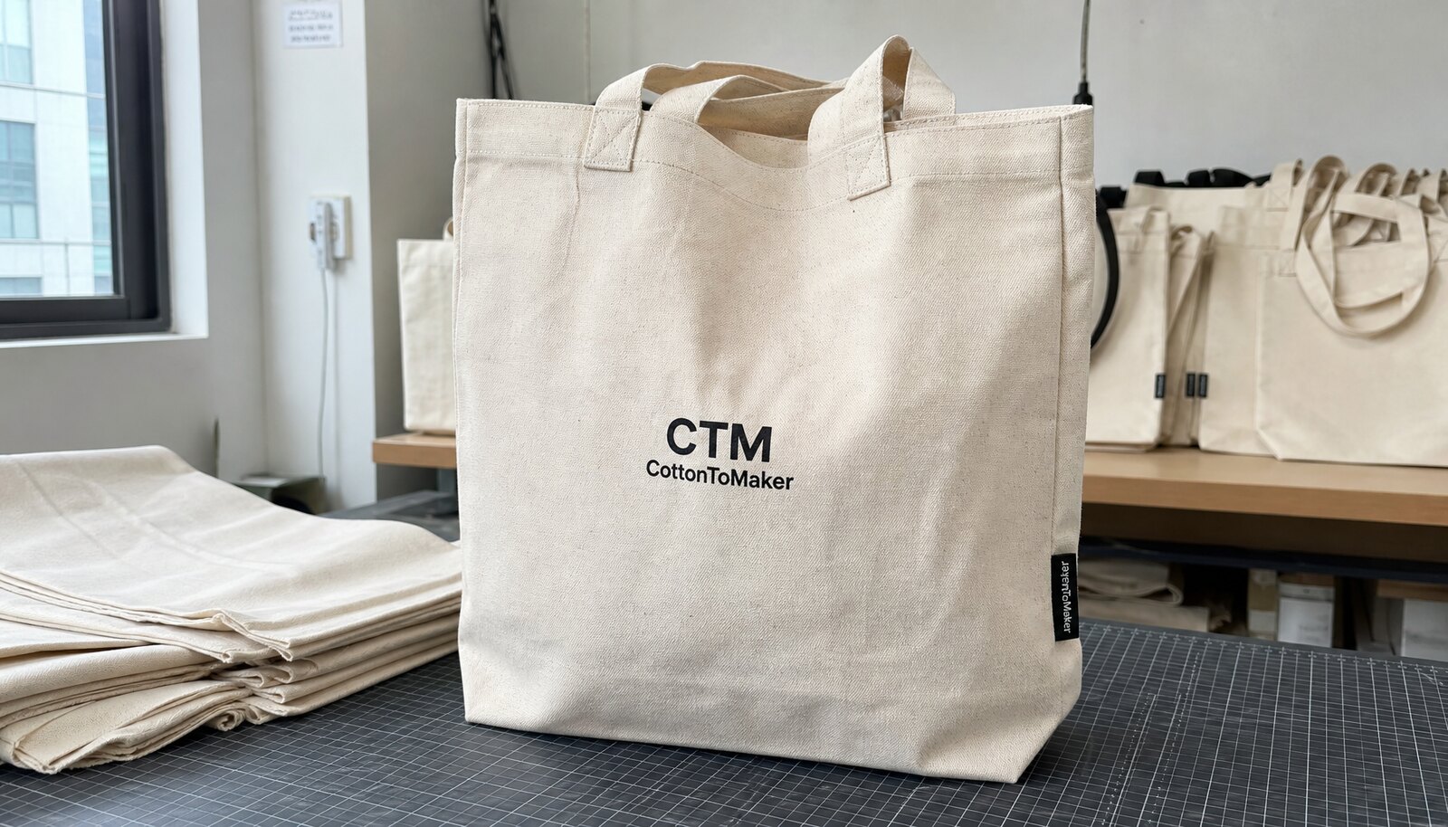 Natural canvas shopping bags with a small CTM CottonToMaker logo print and woven side label in a factory sample room.